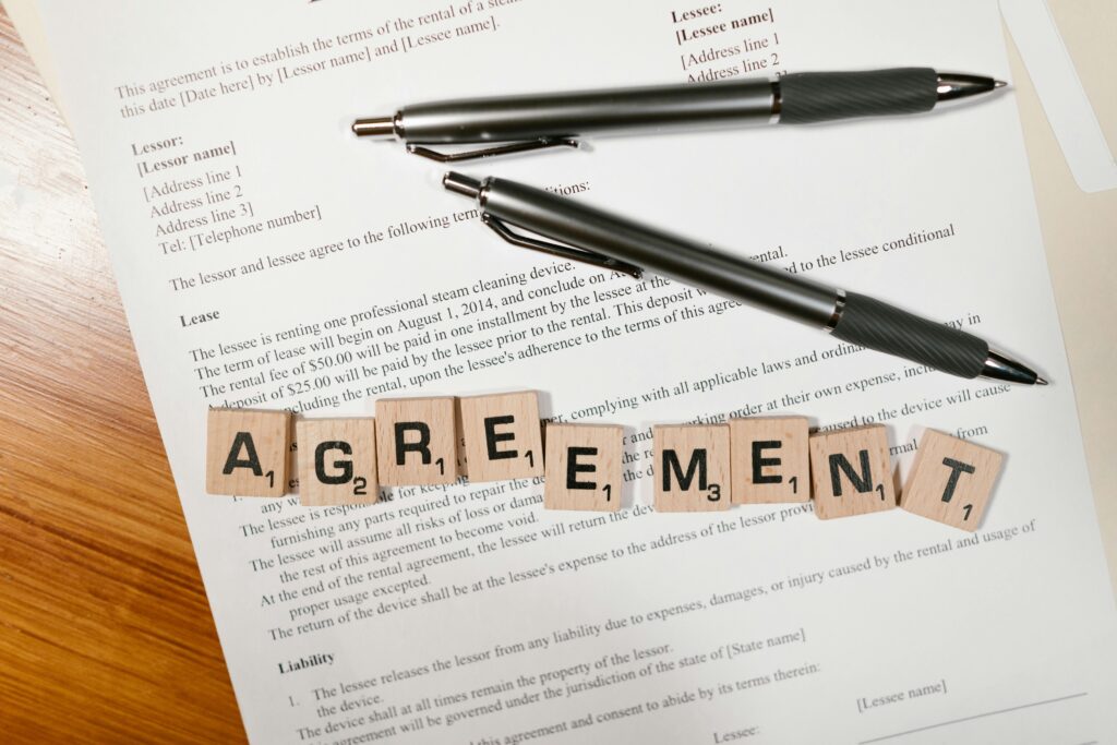 A document with Scrabble tiles spelling 'AGREEMENT' and two pens, symbolizing contract signing.